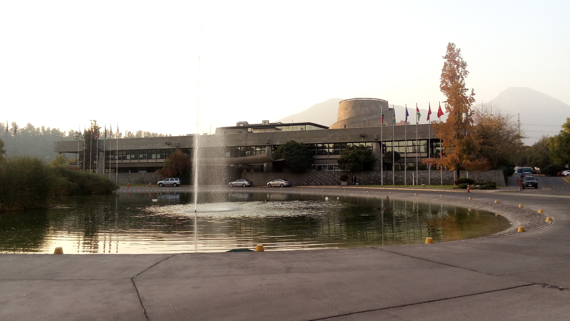 Vista exterior del edificio de la sede de la Comisión Económica para América Latina y el Caribe (CEPAL) con una fuente de agua en primer plano y banderas internacionales de fondo.