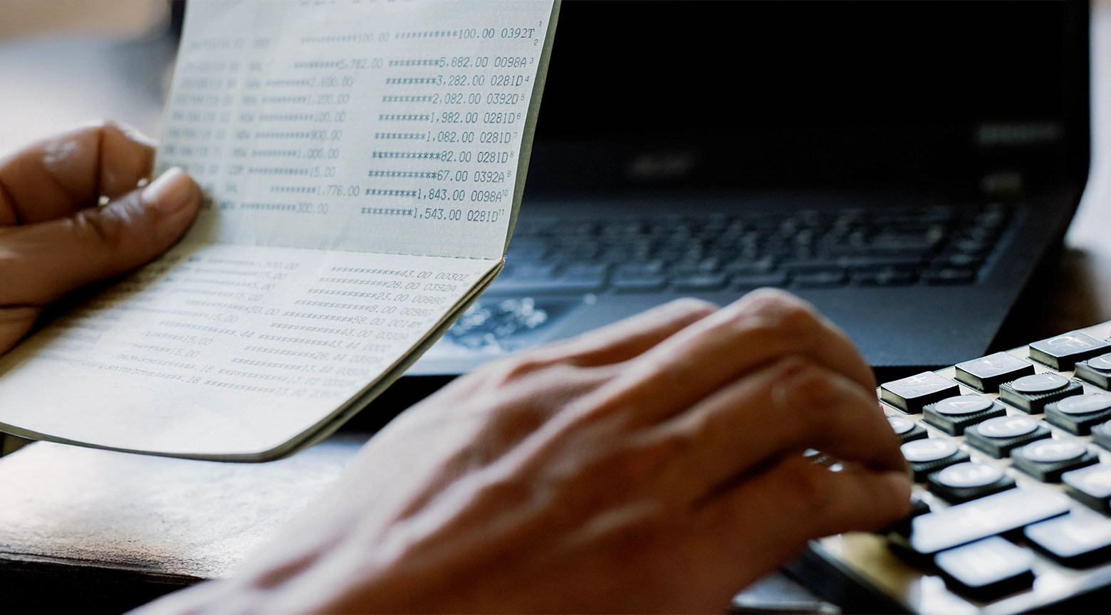 Persona sostiene una libreta bancaria con registros de movimientos y cantidades de dinero mientras utiliza una calculadora, con una computadora portátil al fondo.