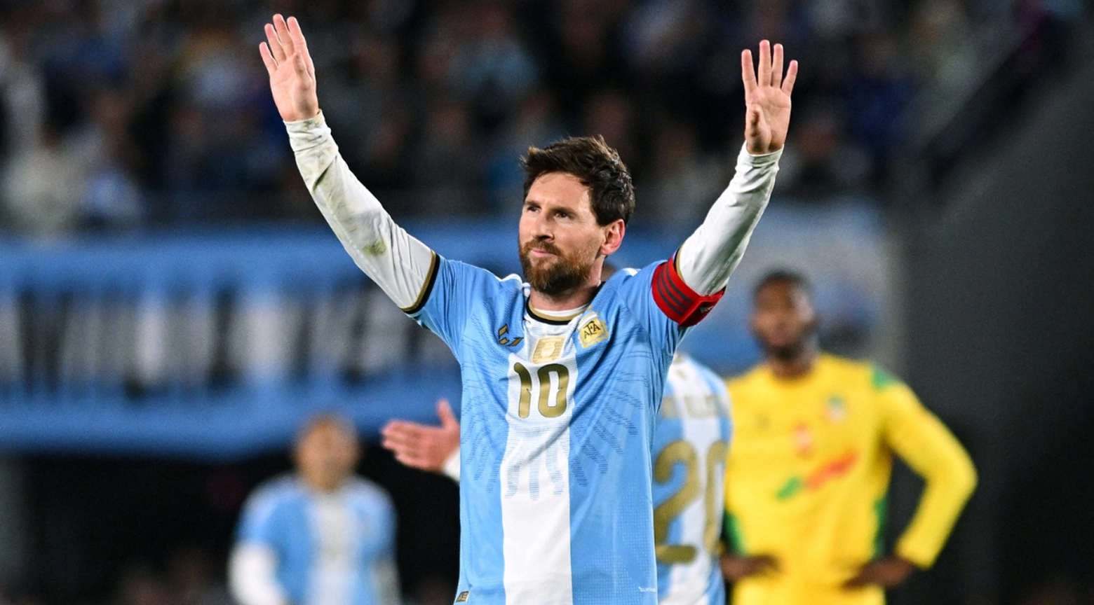 Lionel Messi, con la camiseta de la selección argentina y el brazalete de capitán, saluda a los aficionados con ambos brazos levantados durante un partido.