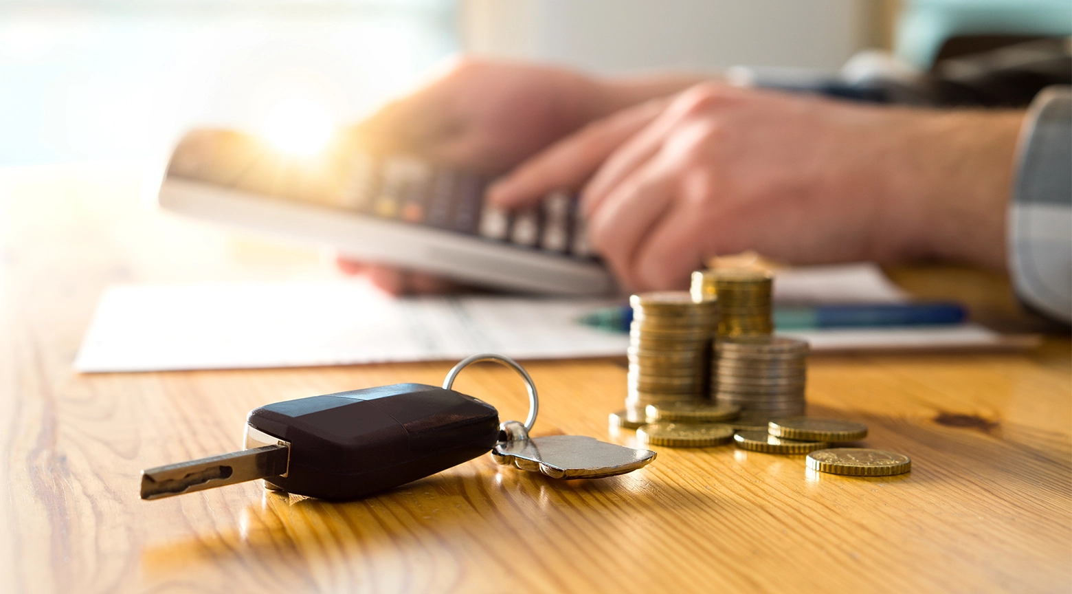 Llave de automóvil y monedas apiladas sobre una mesa de madera con una persona calculando gastos al fondo.