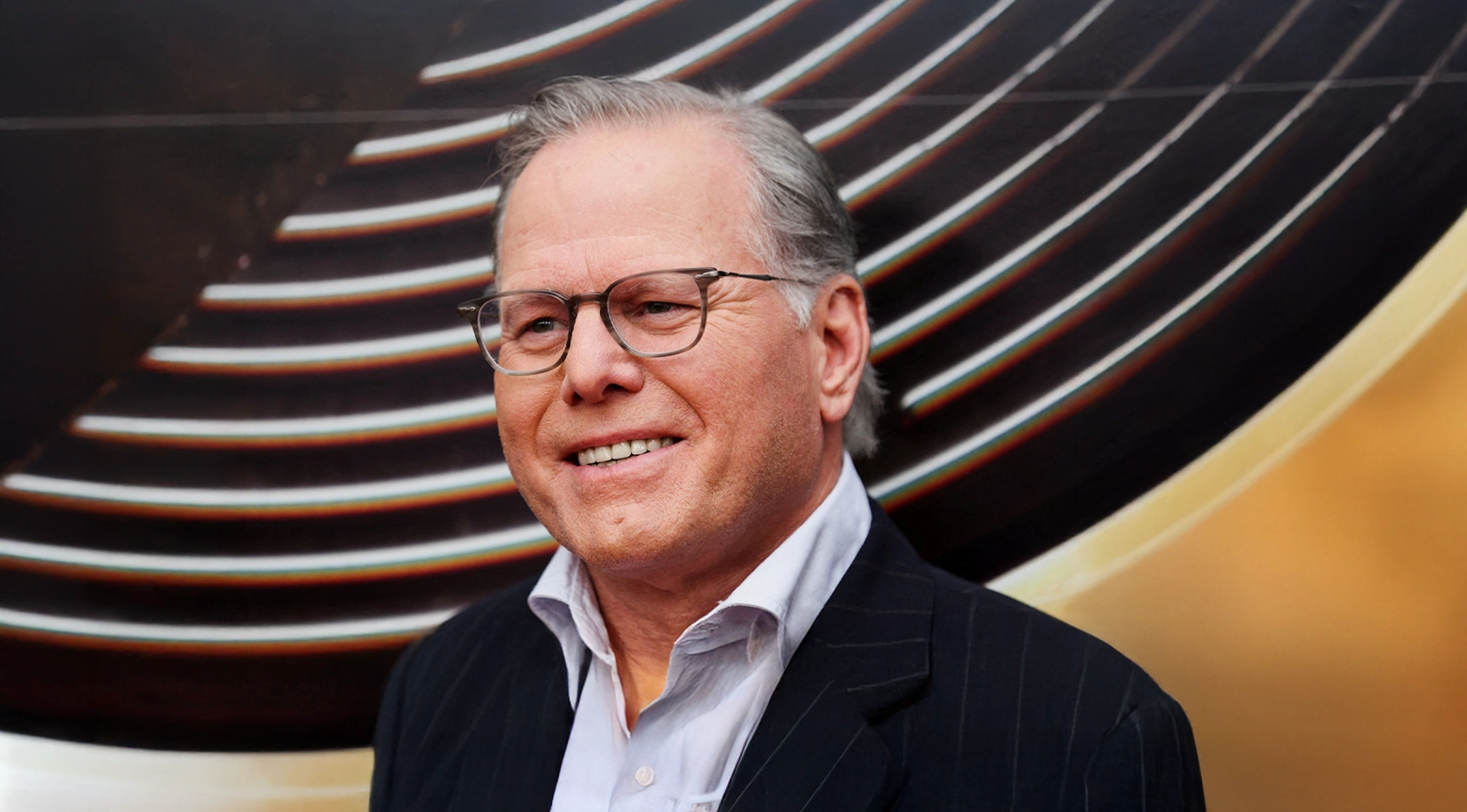 Retrato de un ejecutivo de cabello gris y gafas, vestido con traje oscuro y camisa clara, sonriente ante un fondo negro con líneas curvas iluminadas.