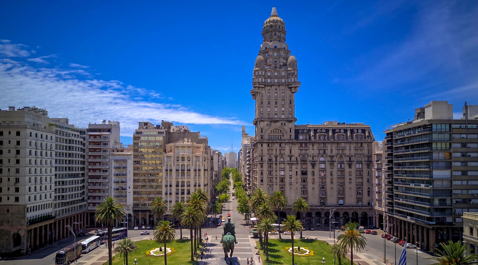 Vista panorámica de la Plaza Independencia de Montevideo, Uruguay, con el histórico Palacio Salvo, el monumento ecuestre a José Gervasio Artigas, jardines, palmeras y edificios urbanos bajo un cielo azul.