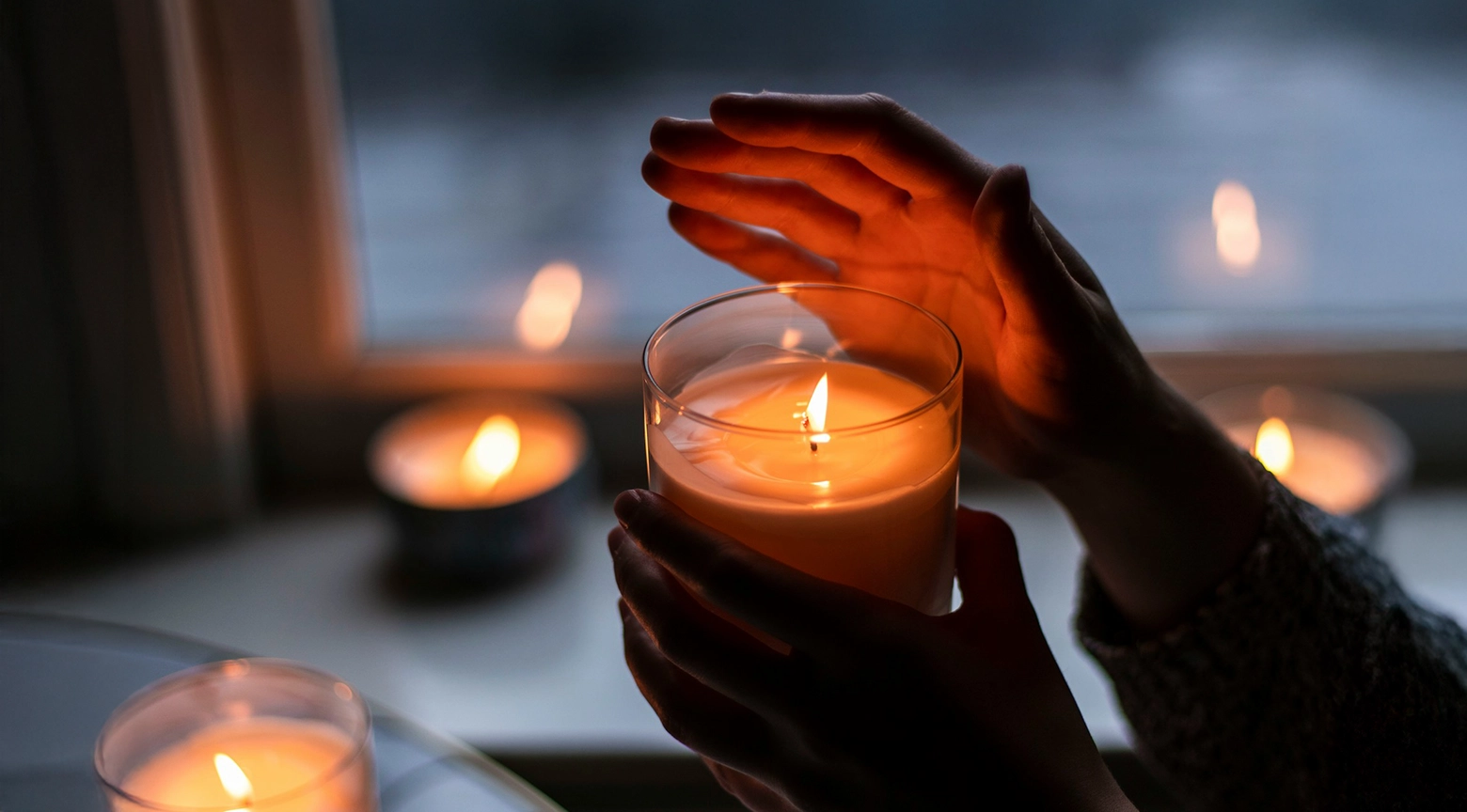 Persona sosteniendo una vela encendida entre sus manos, rodeada de otras velas iluminadas en un ambiente oscuro, como representación de la falta de electricidad.